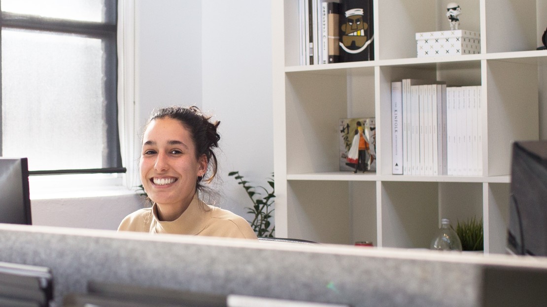Eine junge Frau sitzt an einem Büroarbeitsplatz und lächelt in die Kamera.